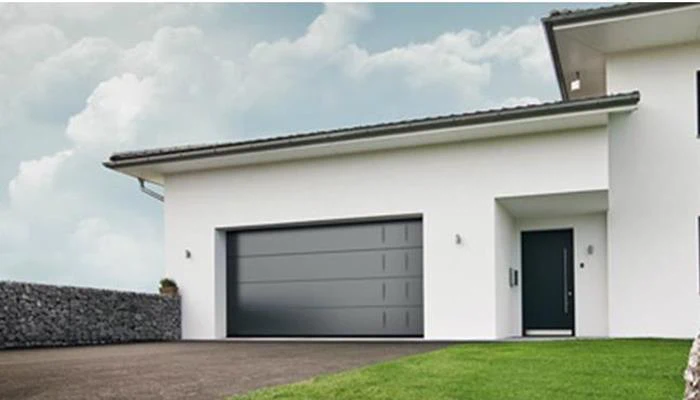 Moderne hvidt hus med en stor grå garageport, sort hoveddør, skrånende indkørsel og grøn græsplæne under en overskyet himmel.