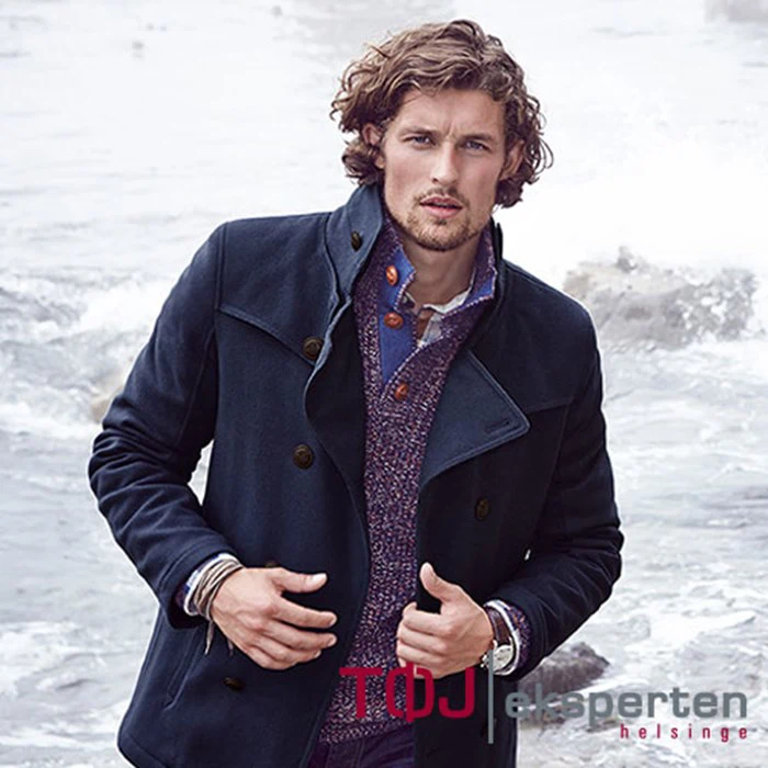 En mand med krøllet hår iført marineblå frakke og lilla sweater står ved klippekysten og kigger ind i kameraet. Ordene TDJ eksperten helsinge står i nederste højre hjørne.