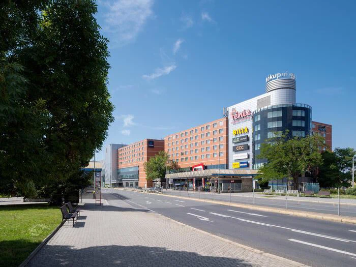 En bred gade med fortov og bænke løber ved siden af et moderne indkøbs- og forretningscenter med forskellige butiksskilte under en klar, blå himmel.