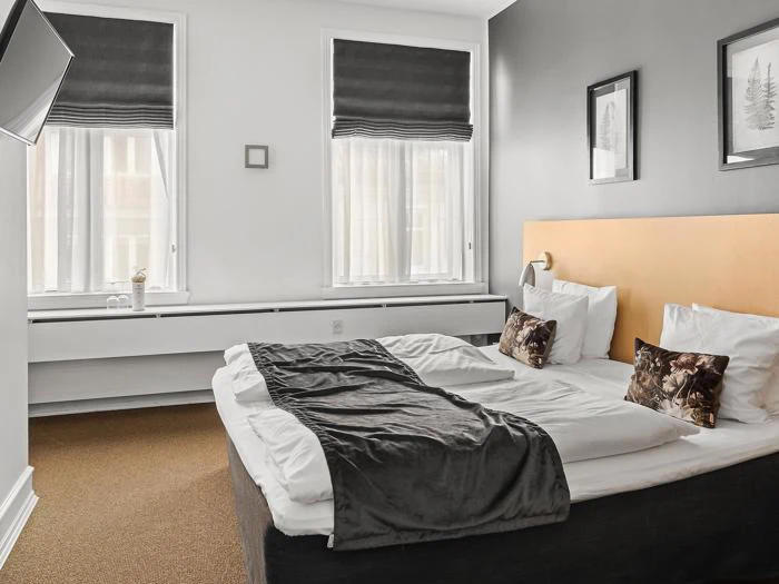 Et moderne hotelværelse med en stor dobbeltseng, et gråt tæppe, to vinduer med mørke persienner, indrammet kunst over sengen og et væghængt tv.