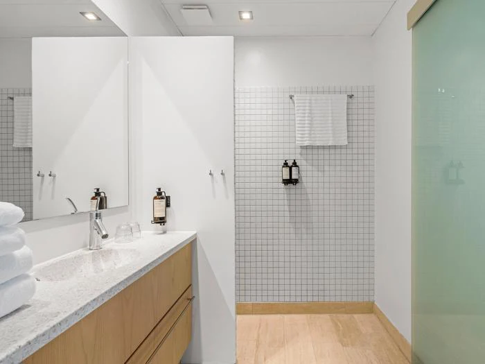 Moderne badeværelse med vaskeskab i lyst træ, stablede hvide håndklæder, væghængt sæbe, flisevæg, håndklædeholder og en dør i matteret glas.