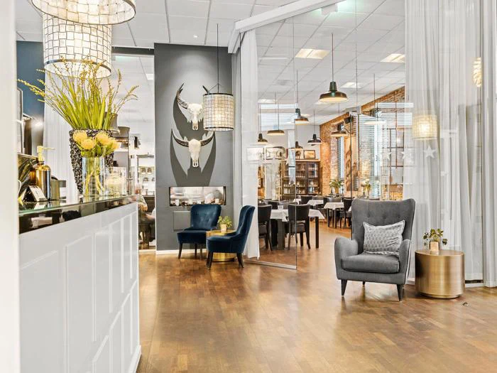 Moderne, stilfuld restaurantindretning med trægulve, elegante siddepladser, dekorative tyrekranier på en grå væg og store hængelamper. En reception og et hyggeligt loungeområde er synlige.