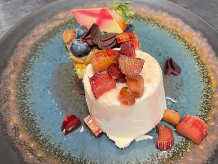 En anrettet dessert består af en cylinder af cremet hvid mousse toppet med rabarber i tern, ved siden af en lille tærte pyntet med blåbær og et dekorativt stykke chokolade på en blå keramiktallerken.