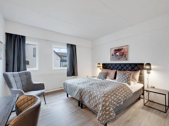 Moderne soveværelse med en dobbeltseng, mønstret sengetøj, to sengelamper, en grå lænestol, et skrivebord med en stol, to vinduer og vægkunst over sengen.