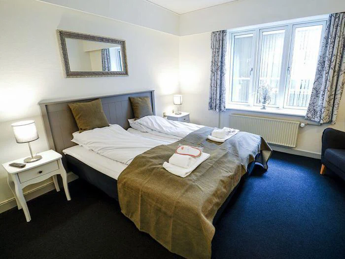 Et pænt hotelværelse med en dobbeltseng, natborde med lamper, et stort spejl over sengen og et vindue med gardiner, der lukker naturligt lys ind. Håndklæderne ligger pænt på sengen.