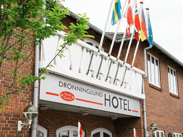 Indgang til Dronninglund Hotel med flere flag over, et træ til venstre og en murstensfacade i baggrunden.