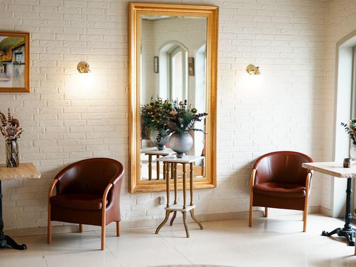 To brune stole står over for et stort spejl med guldramme på en hvid murstensvæg. En vase med blomster står på et lille bord mellem stolene, og der er væglamper over hver af dem.