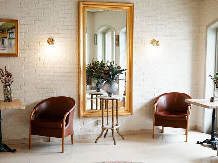 To brune stole står over for hinanden ved siden af et stort guldindrammet spejl på en hvid murstensvæg, med små borde og vaser med blomster imellem og ved siden af dem.