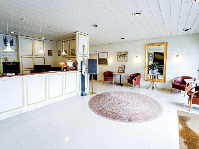 En lys hotellobby med hvid receptionsskranke, rundt tæppe, brune stole, vægkunst, et stort spejl og hvidt flisegulv.