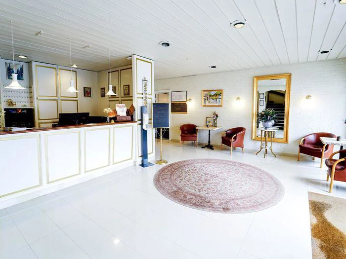 Lys hotellobby med hvid reception, røde stole, et rundt tæppe, et stort vægspejl og indrammede kunstværker på hvide vægge.