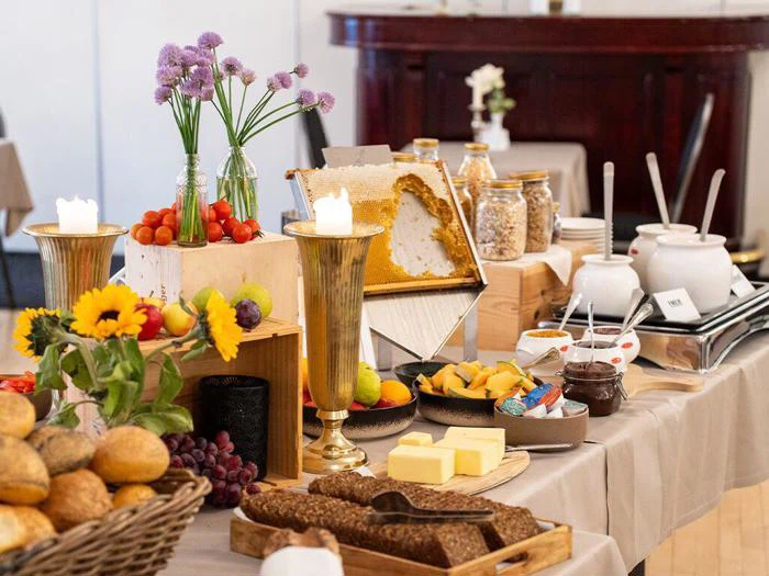 Et buffetbord med brød, ost, frugt, honningkage, morgenmadsprodukter, smør, stearinlys og friske blomster i vaser arrangeret på beige duge.