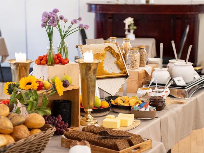 En morgenmadsbuffet med brød, ost, frugt, pålæg, honningkage, cornflakes, blomster og stearinlys på et bord dækket med en beige dug.
