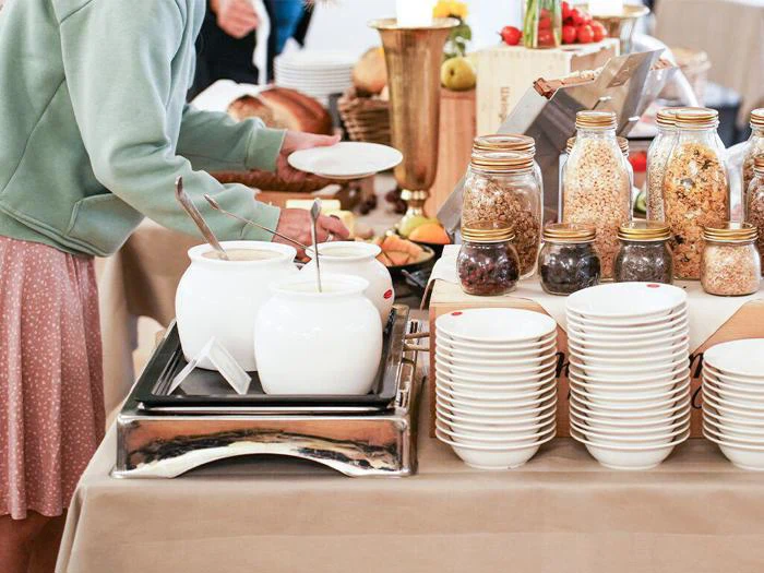 En person i en grøn sweater vælger mad ved et buffetbord med krukker med morgenmadsprodukter, frugt, skåle og suppebeholdere.
