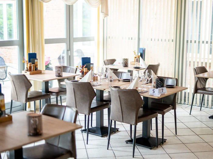 Et lyst, moderne restaurantområde med pænt dækkede borde, brune stole, menuer, glas og naturligt lys, der strømmer ind gennem store vinduer.