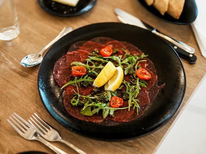 En sort tallerken med tynde skiver rå oksecarpaccio, toppet med rucola, cherrytomater og to citronbåde, på et træbord med bestik og brød i nærheden.