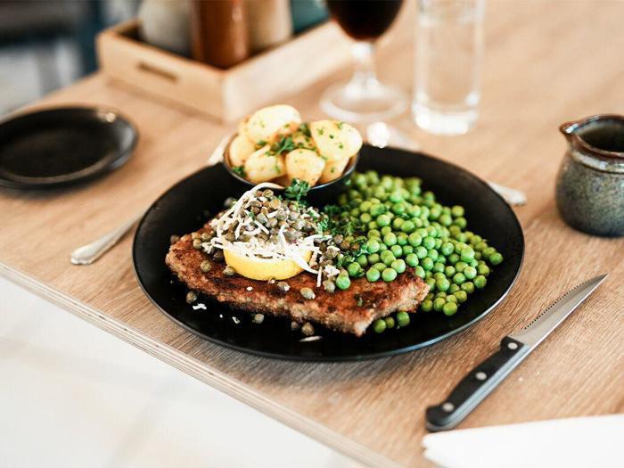 En tallerken med paneret kød toppet med citron, kapers og sauce, serveret med grønne ærter og ristede kartofler, på et træbord med bestik og drikkevarer.