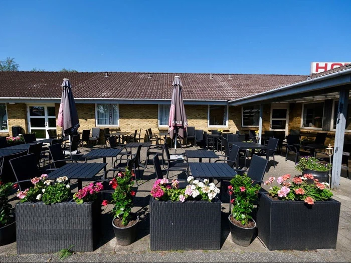 Udendørs terrasse med sorte borde og stole, lyserøde og hvide blomster i potter foran, store parasoller og en murstensbygning i baggrunden under en klar blå himmel.