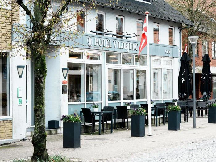 Gadebillede af Hotel Vildbjergs hvide facade med store vinduer, udendørs siddepladser, plantekasser med blomster og et dansk flag ved indgangen.