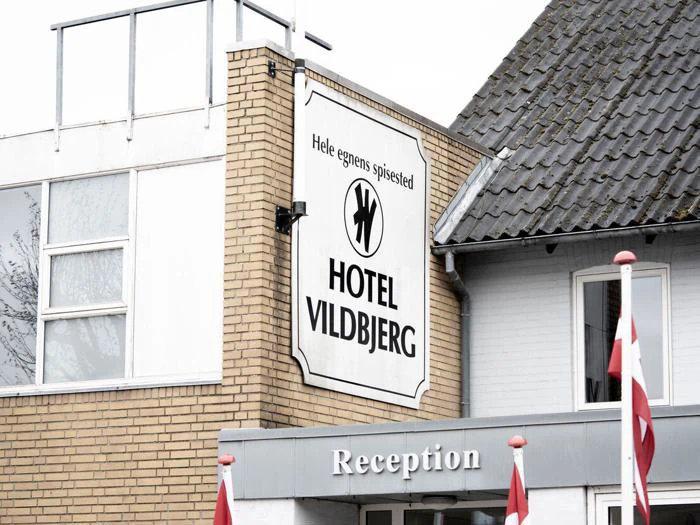 Skilt for Hotel Vildbjerg med dansk tekst på en murstensbygning over en grå receptionsindgang; en del af taget og danske flag er synlige.