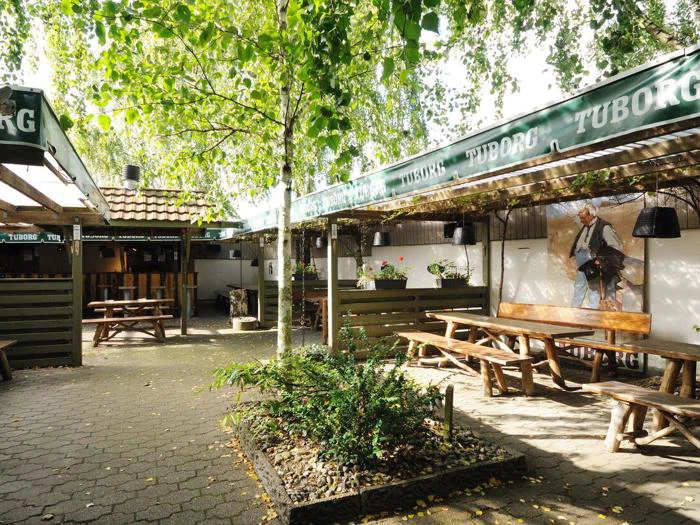 Udendørs siddeområde med træbænke, grønne Tuborg-markiser, grønne træer, der giver skygge, og et vægmaleri af en person på en hvid væg i baggrunden.