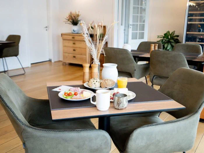 Morgenbord dækket til to med tallerkener med mad, krus, appelsinjuice, brød, salt- og peberbøsse og en hvid kande i en hyggelig, moderne spisestue med grønne stole.