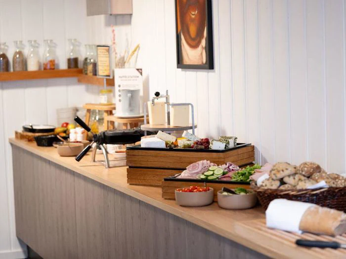 En morgenmadsbuffet med brød, friske grøntsager, ost, pålæg, vaffeljern og krukker med ingredienser på en trædisk mod en hvid væg.