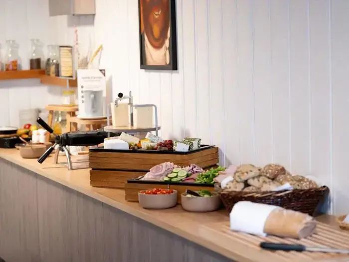 En morgenmadsbuffet på en trædisk med brød, frugt, grøntsager, pålæg, ost og skåle med forskellige toppings i hyggelige omgivelser.