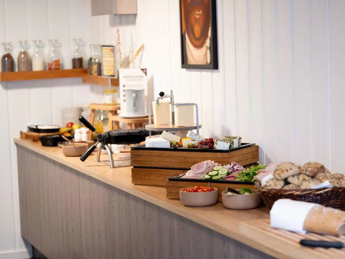 En morgenmadsbuffet med forskelligt brød, ost, kød, grøntsager og krydderier arrangeret på en trædisk i en moderne køkkenindretning.