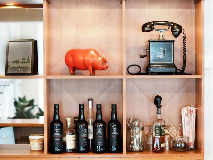 Træhylde med flasker, cocktailredskaber, en krukke med sugerør, en sort vintagetelefon, en rød grisefigur og et indrammet foto.