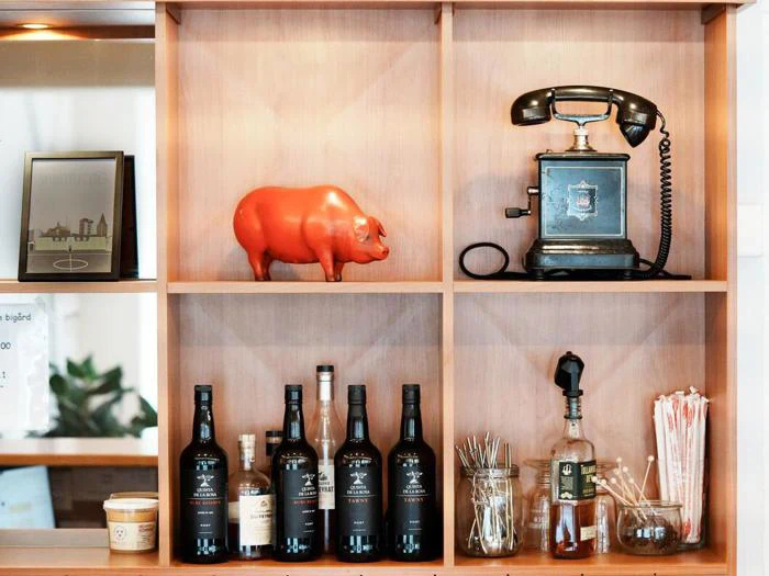 Træhylde med et indrammet foto, en rød grisefigur, en sort vintagetelefon, flasker med alkohol, cocktailpinde, sugerør og en lille skål.