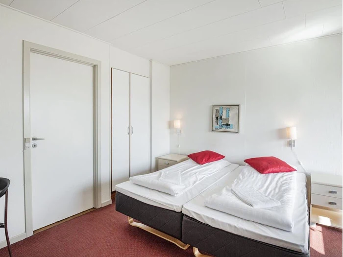 Et simpelt hotelværelse med to enkeltsenge skubbet sammen, hvidt sengetøj, røde puder, natborde, væglamper, et klædeskab og en dør.