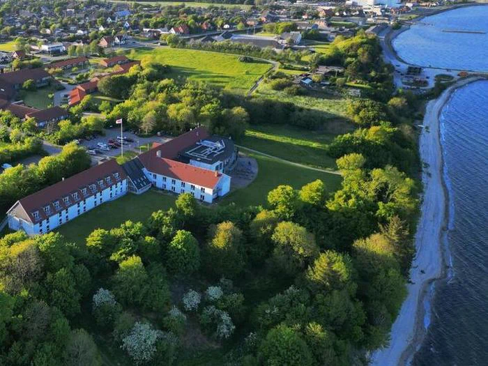 Luftfoto af en stor bygning med rødt tag omgivet af træer nær en kystlinje, med en by og vand i baggrunden.