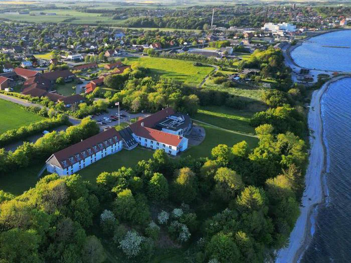 Luftfoto af en stor hvid bygning med rødt tag omgivet af træer nær kysten, med en by og åbne marker i baggrunden.