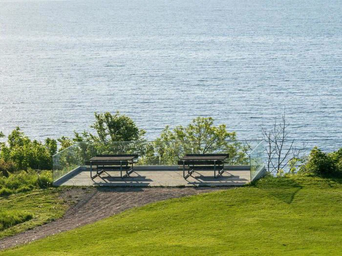 To picnicborde står på et trædæk med glasrækværk med udsigt over et stort vandområde og omgivet af grønt græs og buske.