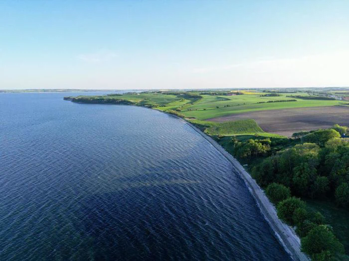 Luftfoto af en kystlinje med roligt blåt vand, grønne marker og træer langs kysten under en klar himmel.