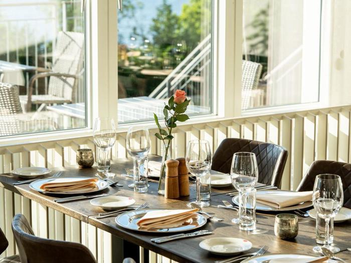 Et solbeskinnet restaurantbord dækket med tallerkener, glas, servietter og en enkelt lyserød rose i en vase ved siden af store vinduer med udsigt til en lys udendørs gårdhave.