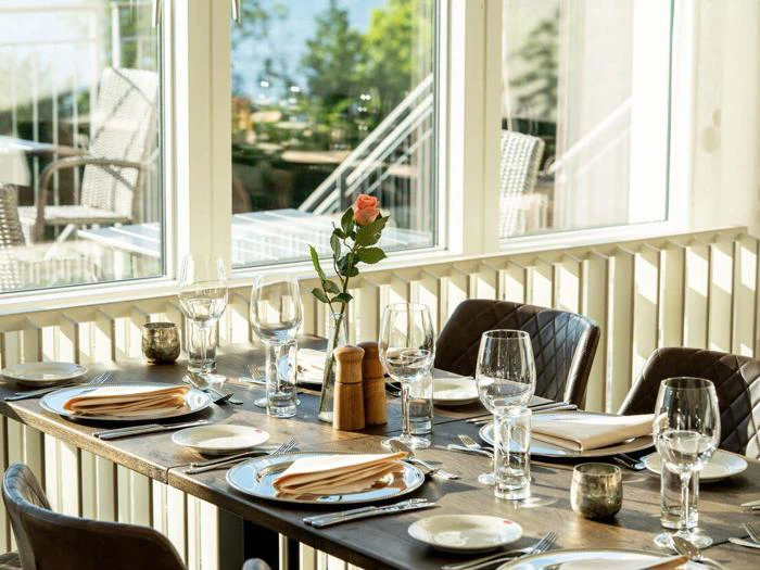 Et solbeskinnet restaurantbord dækket til fire med glas, tallerkener, servietter og en enkelt lyserød rose i en vase ved siden af store vinduer med udsigt til en udendørs terrasse.