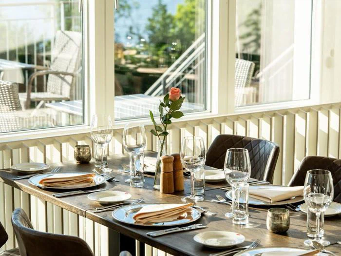 Et solbeskinnet restaurantbord dækket med tallerkener, glas, servietter og en enkelt lyserød rose i en vase ved siden af store vinduer med udsigt til en lys udendørs gårdhave.