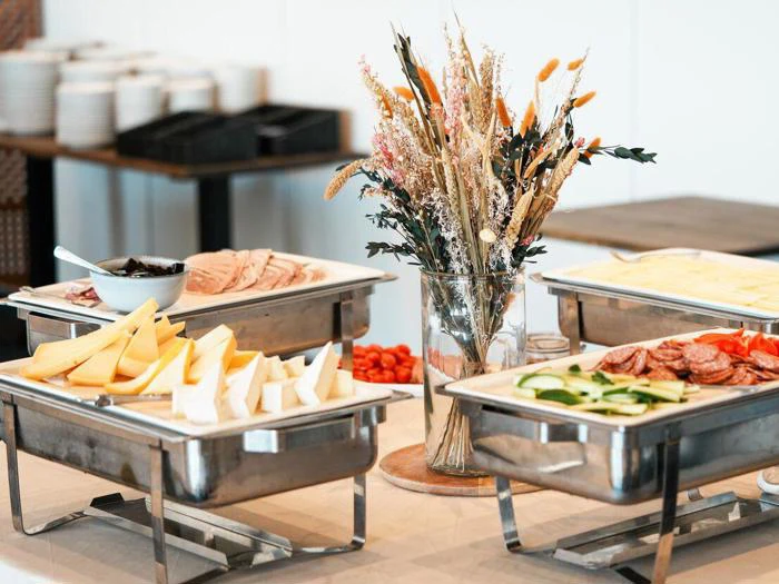 Et buffetbord med bakker med skiveskåret ost, kød, grøntsager og brød med en vase med tørrede blomster som midtpunkt. Tallerkener og kopper er stablet i baggrunden.