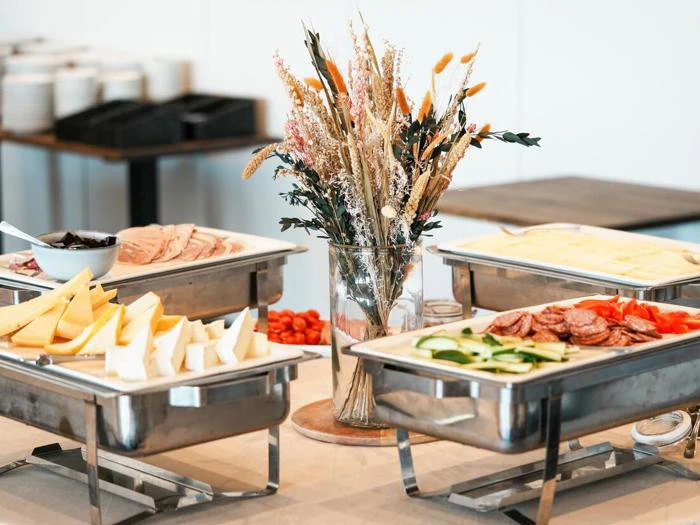 Buffetbord med forskelligt kød, oste, grøntsager og en vase med tørrede blomster som midtpunkt. Tallerkener og bestik er arrangeret i baggrunden.