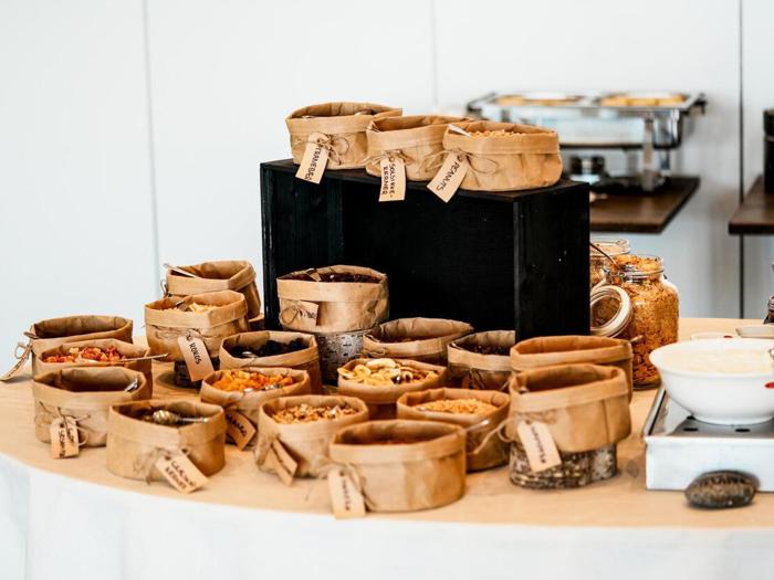 En række mærkede papirposer fyldt med forskellige nødder, frø og tørrede frugter er arrangeret på et bord, sandsynligvis til en morgenmadsbuffet eller snackbar.