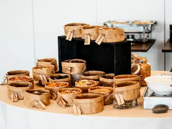 En række mærkede papirposer fyldt med forskellige nødder, frø og tørrede frugter er arrangeret på et bord, sandsynligvis til en morgenmadsbuffet eller snackbar.
