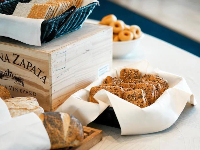 Forskellige brød og kiks i kurve og en trækasse på et bord med en skål med små runde kager i baggrunden.