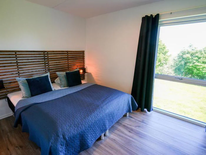 Et moderne soveværelse med dobbeltseng, mørkeblåt sengetøj, hovedgærde af træ og et stort vindue med udsigt til grønne træer og græs.