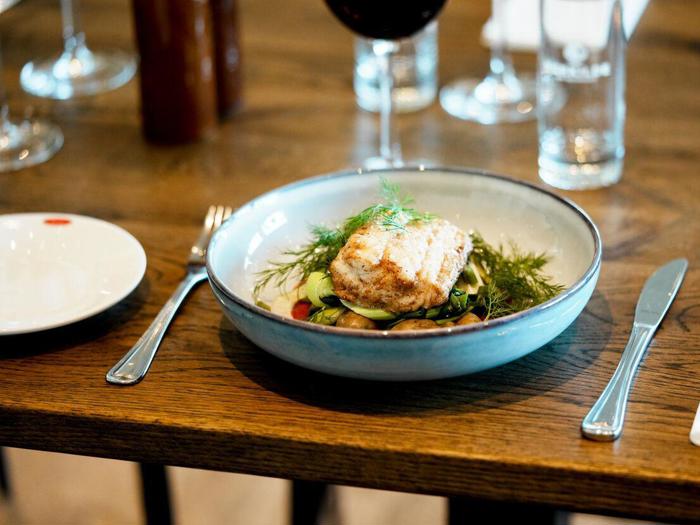 En tallerken med grillet fisk serveret på grøntsager og pyntet med krydderurter står på et dækket spisebord med bestik og glas.
