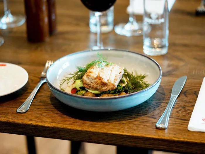 En tallerken med grillet fisk serveret med grøntsager og urter, dækket på et træbord med sølvtøj, et vinglas og et vandglas.