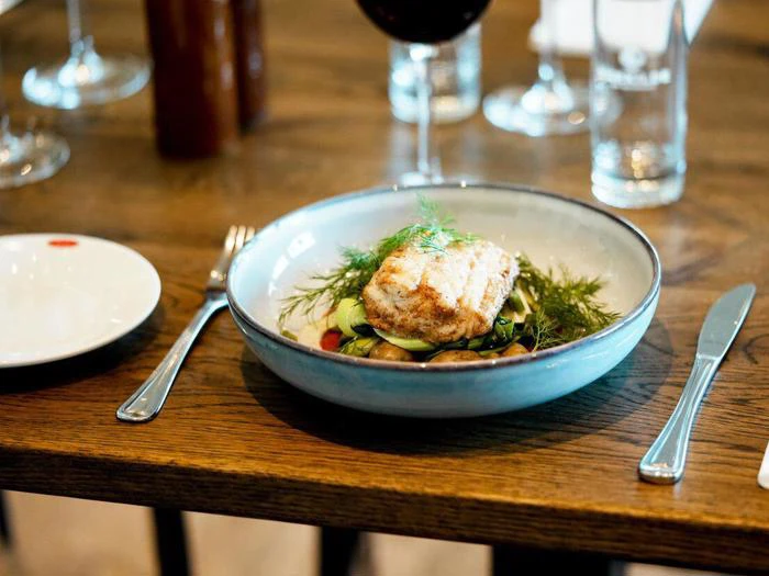 En tallerken med grillet fisk serveret på grøntsager og pyntet med krydderurter står på et dækket spisebord med bestik og glas.