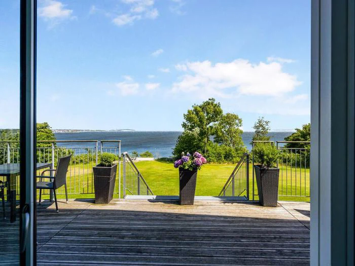 Udsigt fra en træterrasse med potteblomster og havemøbler med udsigt over en græsplæne, træer og et stort vandområde under en blå himmel.