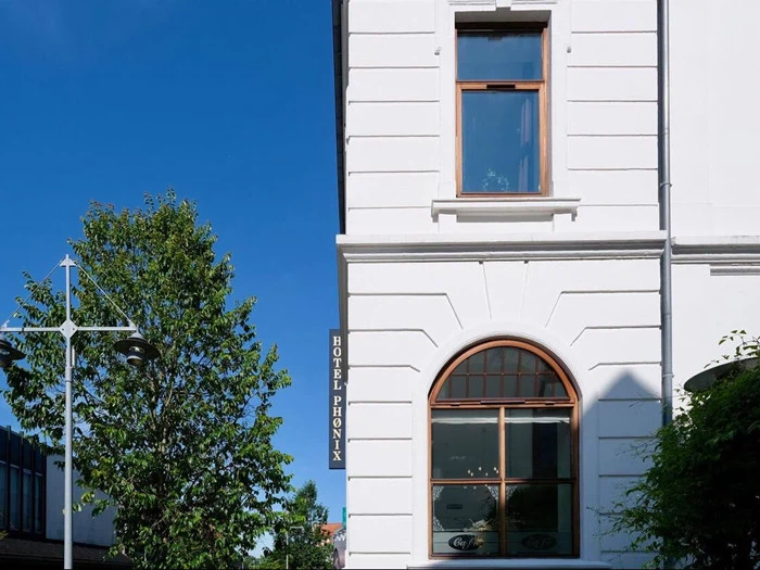 Hvid bygning med træindrammede vinduer og et lodret HOTEL PHOENIX-skilt; grønne træer og en gadelampe er synlige mod en klar blå himmel.