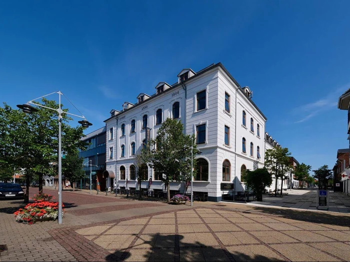 En hvid, treetagers historisk bygning med buede vinduer på et solrigt gadehjørne, omgivet af træer og blomsterbede.